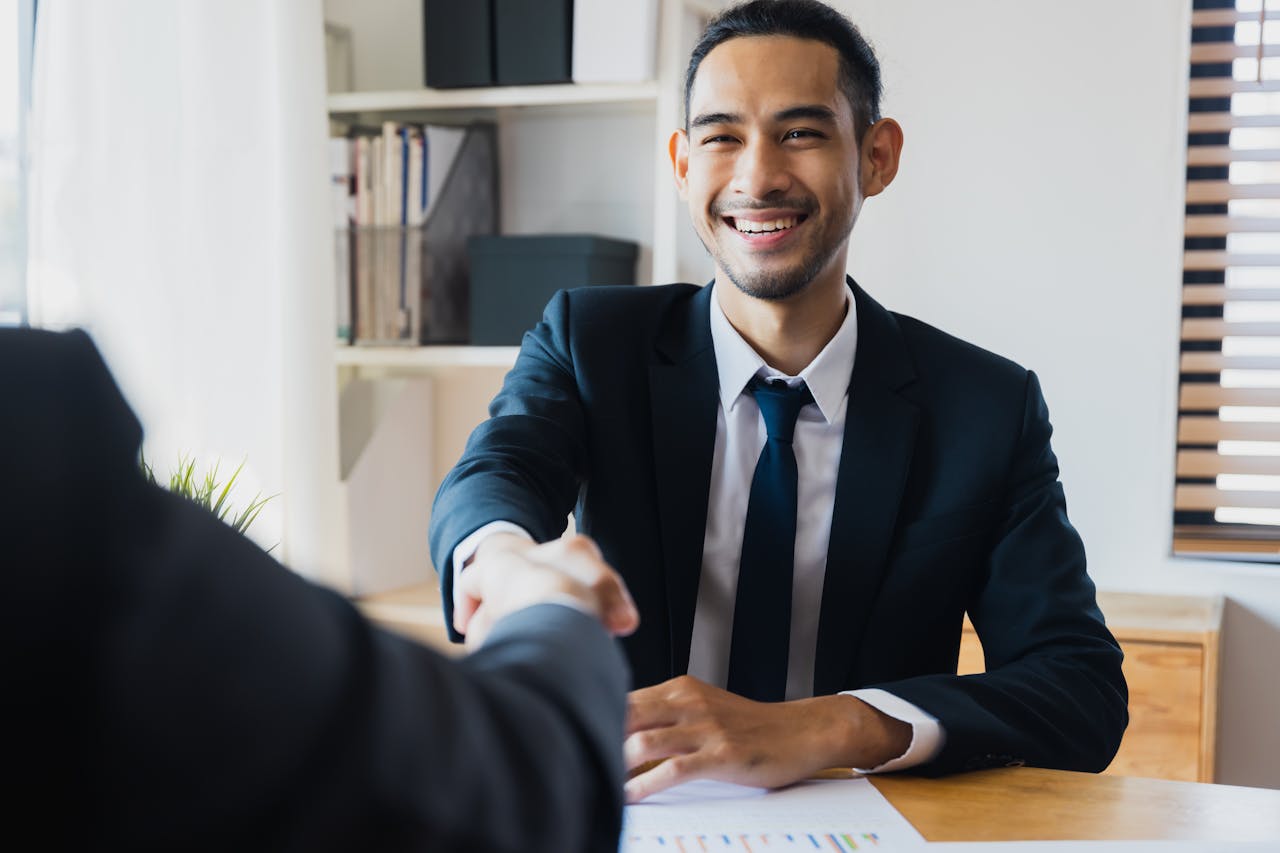 Services-02 Confident businessman in suit shaking hands at office desk, symbolizing successful partnership.